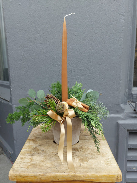 Decorative arrangement with a tall candle, greenery, and a gold ribbon on a wooden table.
