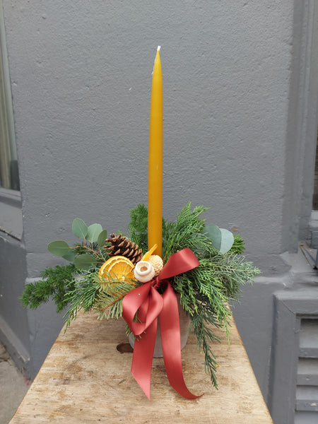 Decorative arrangement with a tall yellow candle, greenery, pine cones, and a red ribbon on a wooden surface.