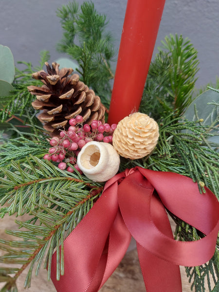 Decorative arrangement with greenery, pine cones, berries, and a red ribbon on a neutral background.