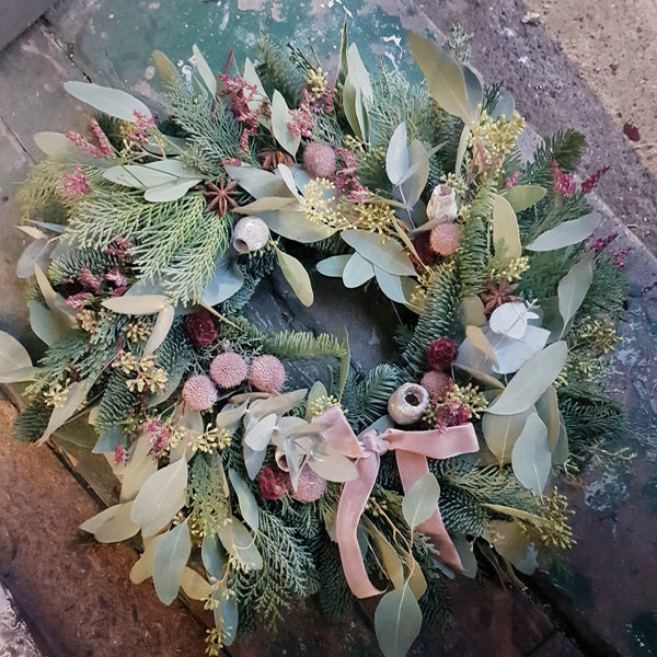 Decorative wreath with greenery, flowers, and a pink ribbon on a textured surface.